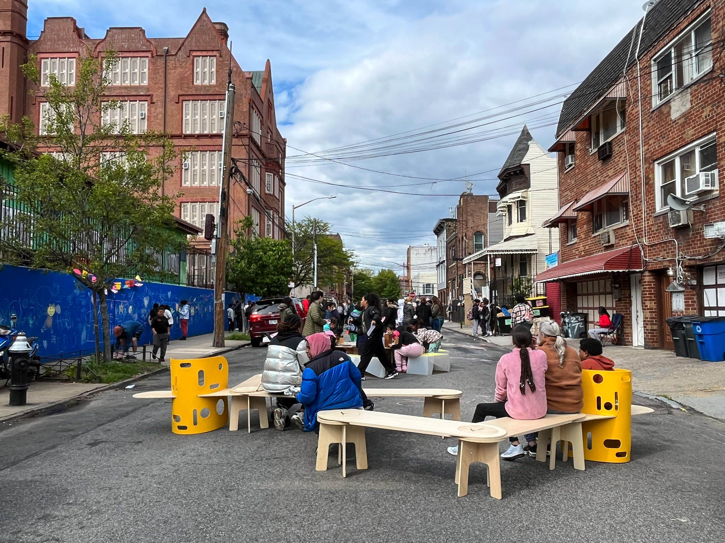 A view of children and adults sitting on a bench on an open street in nyc.